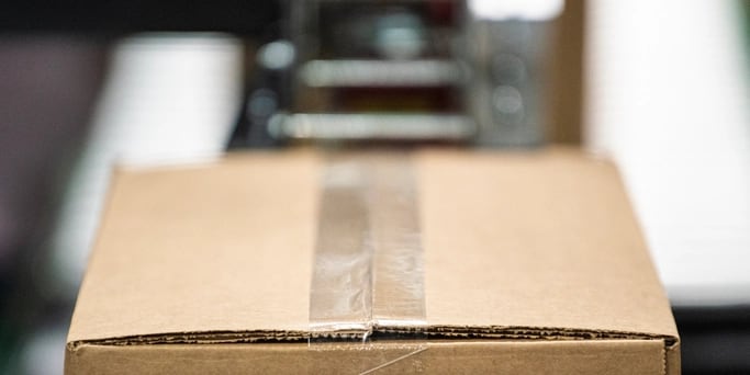 A sealed, taped carton on a packing line with the line falling out of focus behind it.